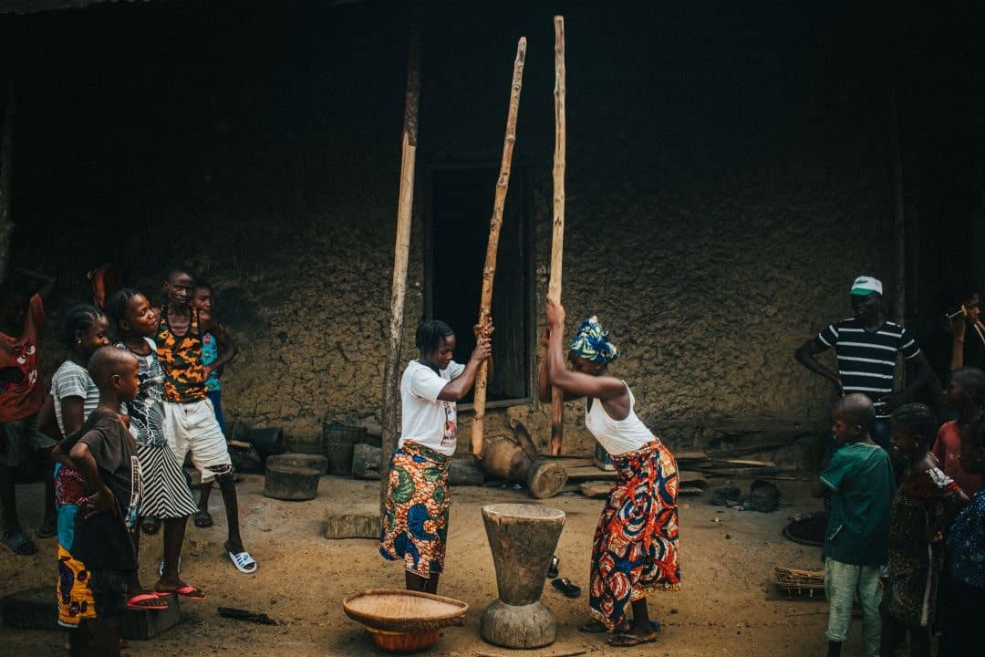 GBL_5-min 2 women wearing colorful skirts pound rice with long wooden sticks