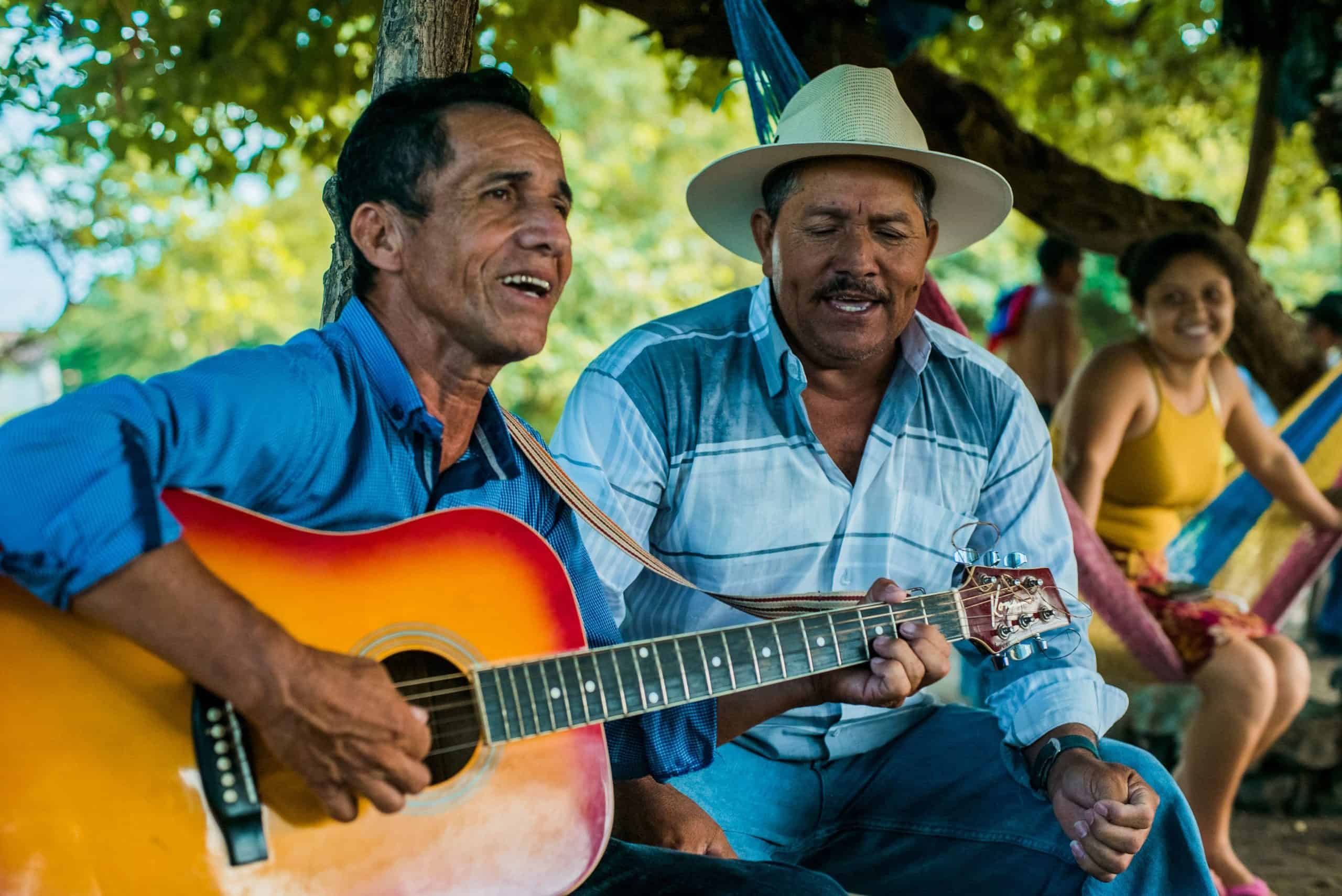 Two men in a rural Honduran community sit side by side at a local protest site, laying the guitar by side