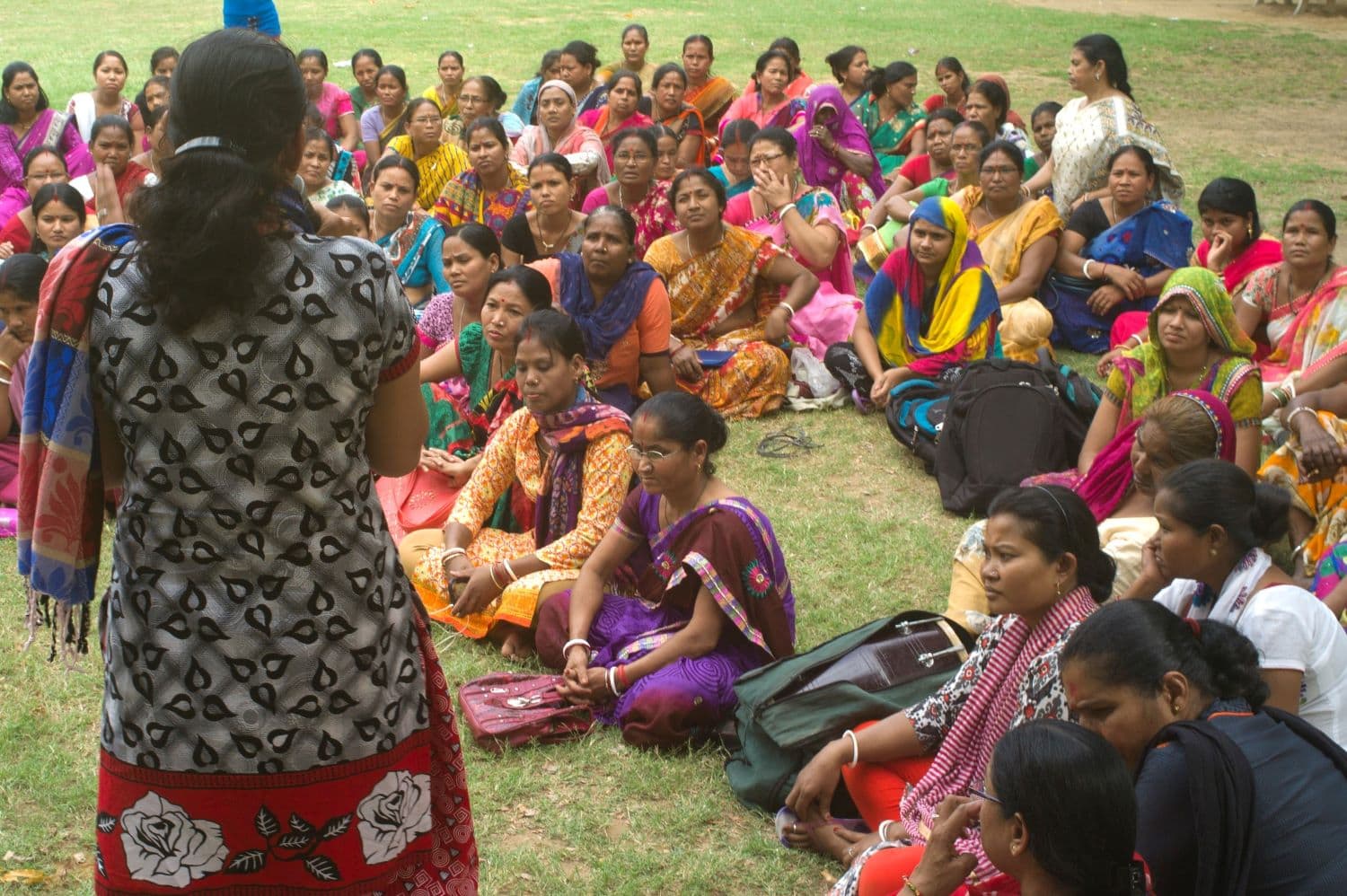 A group of women sit in a park listening to a speaker in a printed black and white sari