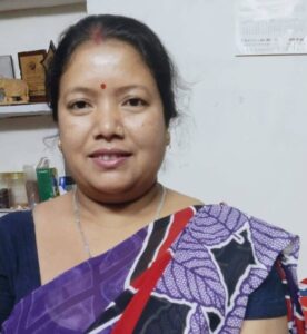 A young woman with a bindi and blue and purple patterned sari stands in front of a bookcase and smiles