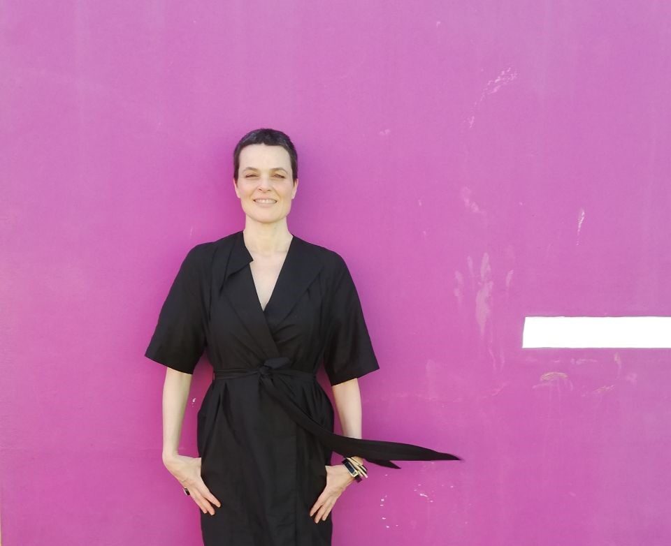 A person in a black dress stands in front of a pink wall.