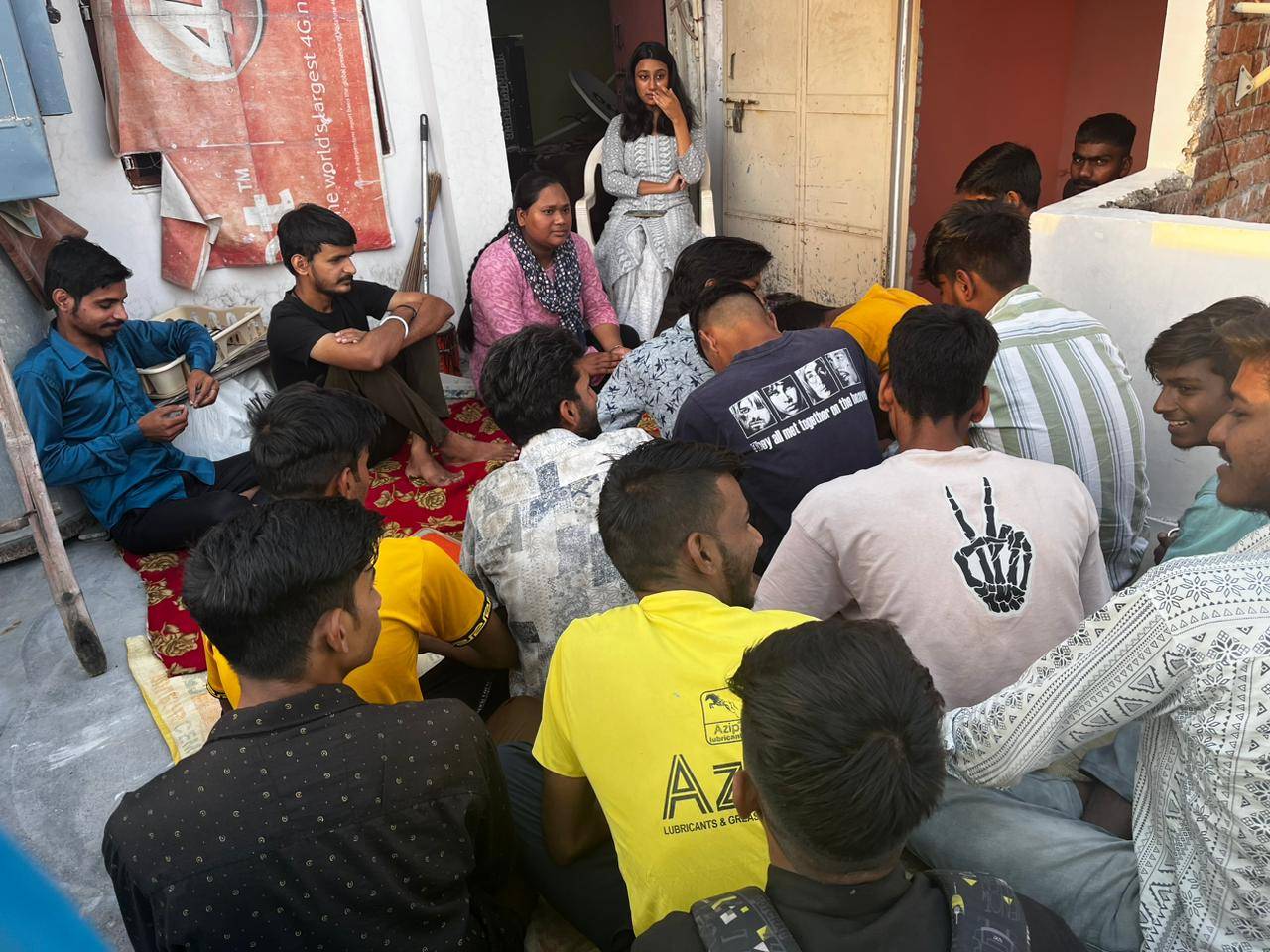 A group of men in their early 20s sit in front of two women at an informal outdoor workshop.