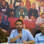 Three men sit at a table under a mural giving a press conference.