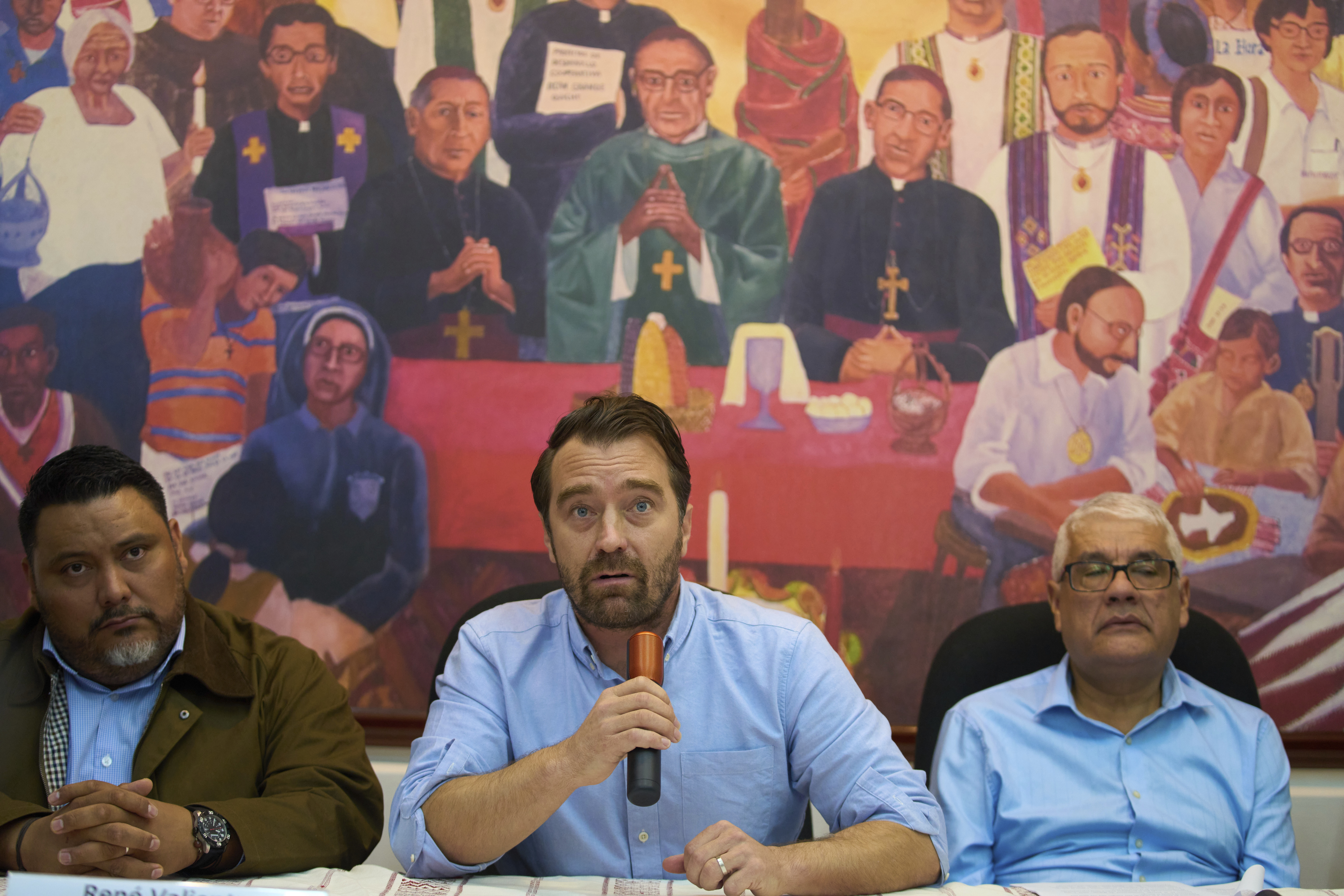 Three men sit at a table under a mural giving a press conference.