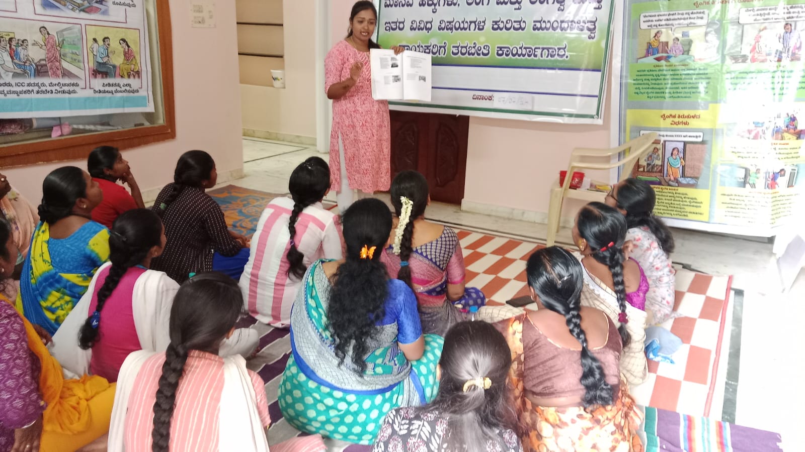 KOLAR (14) Women garment workers sit on the floor in front of a woman who is holding up a book and giving a presentation.