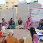 Women sit in a circle on the ground outside in front of a banner that reads "Rural Environment Awareness Community Help (REACH)."