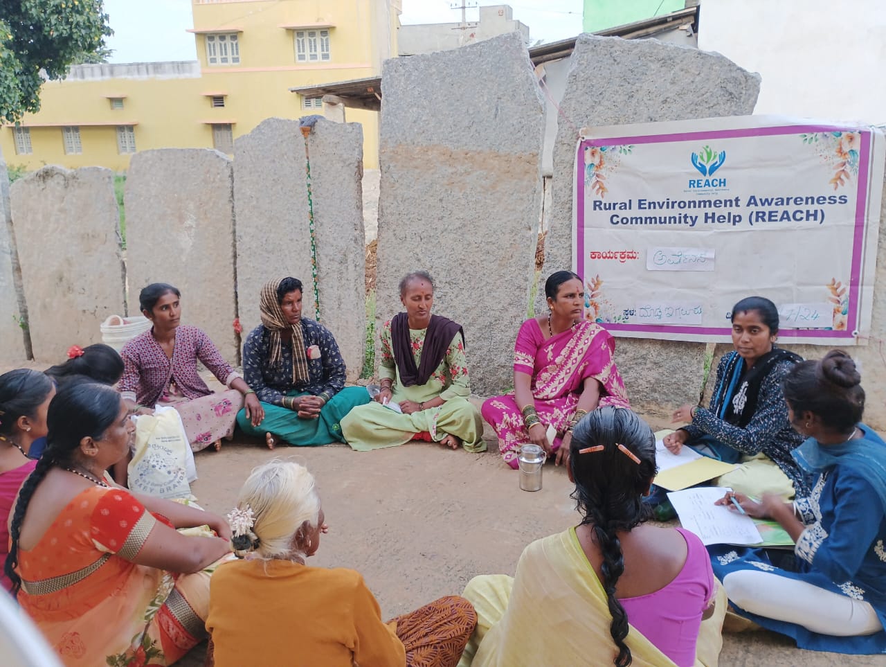 KOLAR (9) Women sit in a circle on the ground outside in front of a banner that reads "Rural Environment Awareness Community Help (REACH)."